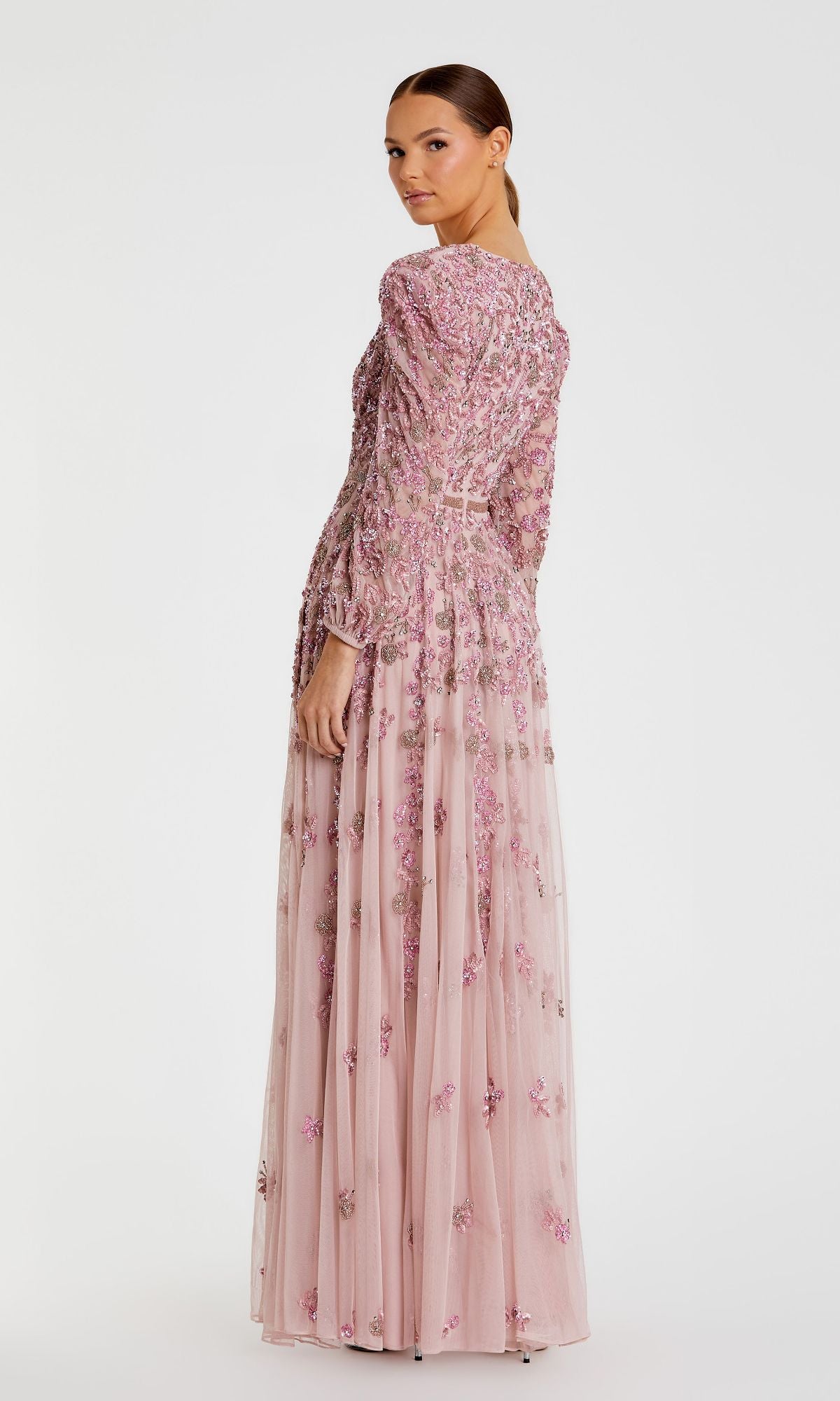 A woman stands facing backward, looking over her shoulder. She is wearing the Mac Duggal 5497—a modest, long-sleeve beaded formal dress in light pink with floral embroidery.