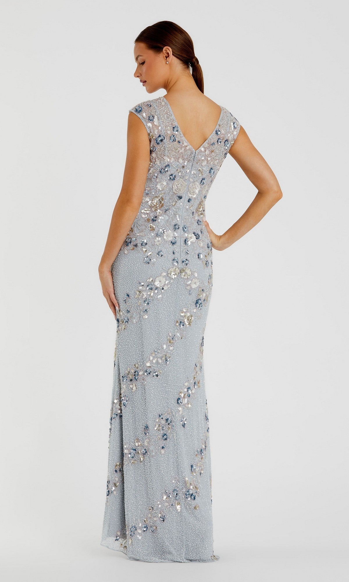 A woman faces away in the Mac Duggal 5500 Beaded Long Formal Dress, a light blue floor-length gown featuring cap sleeves, a V-shaped back, and shimmering beadwork against a plain light background.