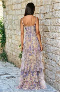 A woman with long brown hair wears the Blondie Nites 5500BN Floral-Print Long Prom Dress with thin straps, standing on a stone path by a wall, facing away from the camera.