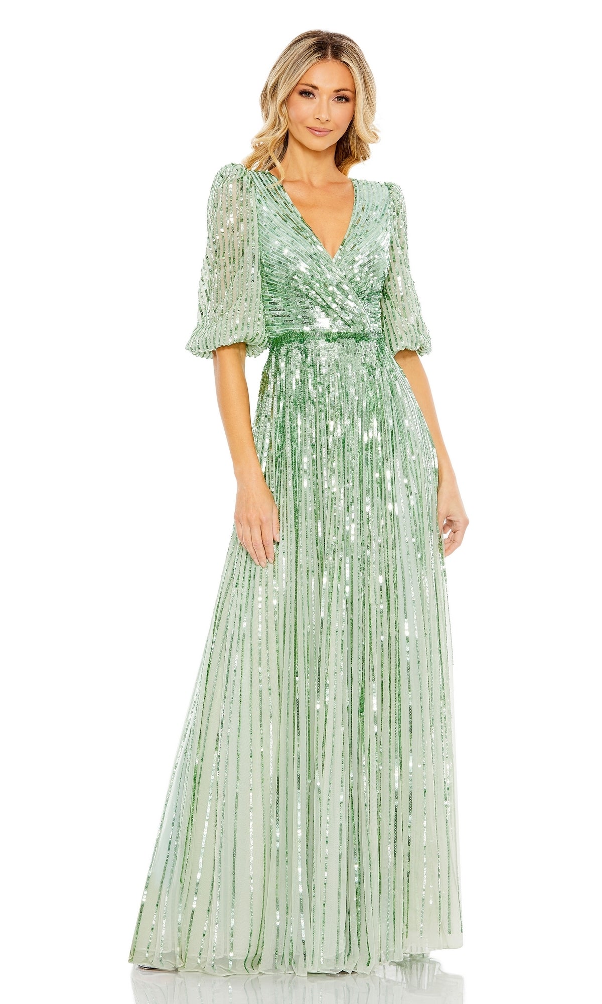 A woman wears the Mac Duggal 5509 long sleeve, floor-length formal dress in light green with sequins, sheer sleeves, a V-neckline, and pleated skirt as she stands against a white background.