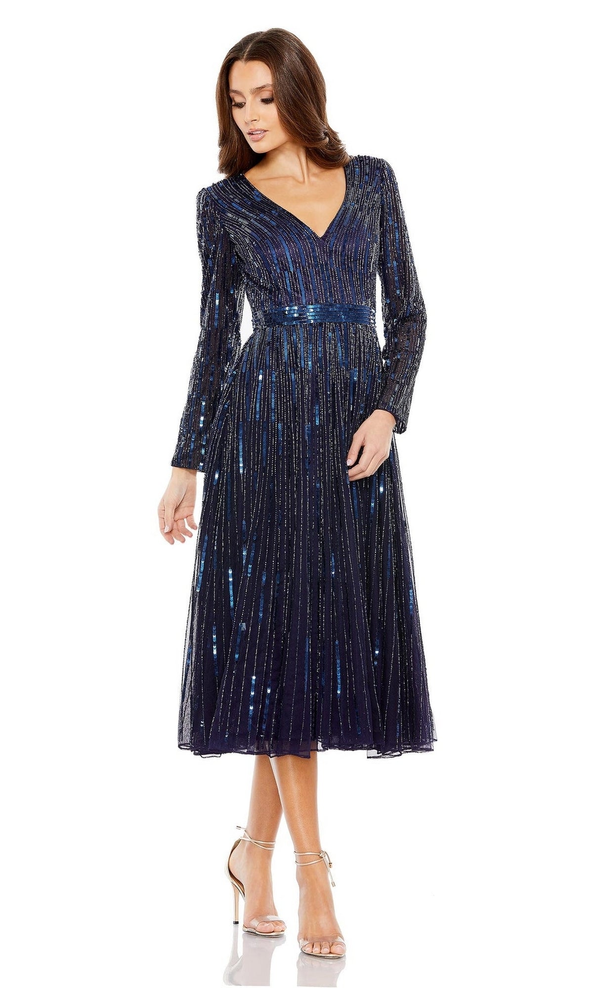 A woman wearing the Short Semi-Formal Dress 5528 by Mac Duggal stands with one leg slightly forward, looking down; the dress features a navy blue color, long sleeves, a V-neck, sequins, and a belted waist.