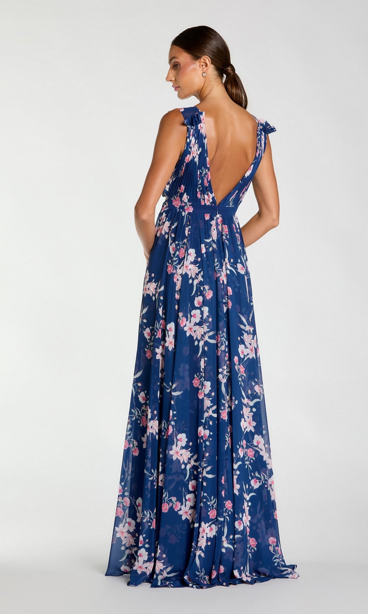 A woman in a Classic Long Formal Dress: Mac Duggal 55321, featuring a blue floral Bohemian design and deep V-back, stands facing away from the camera against a plain background.