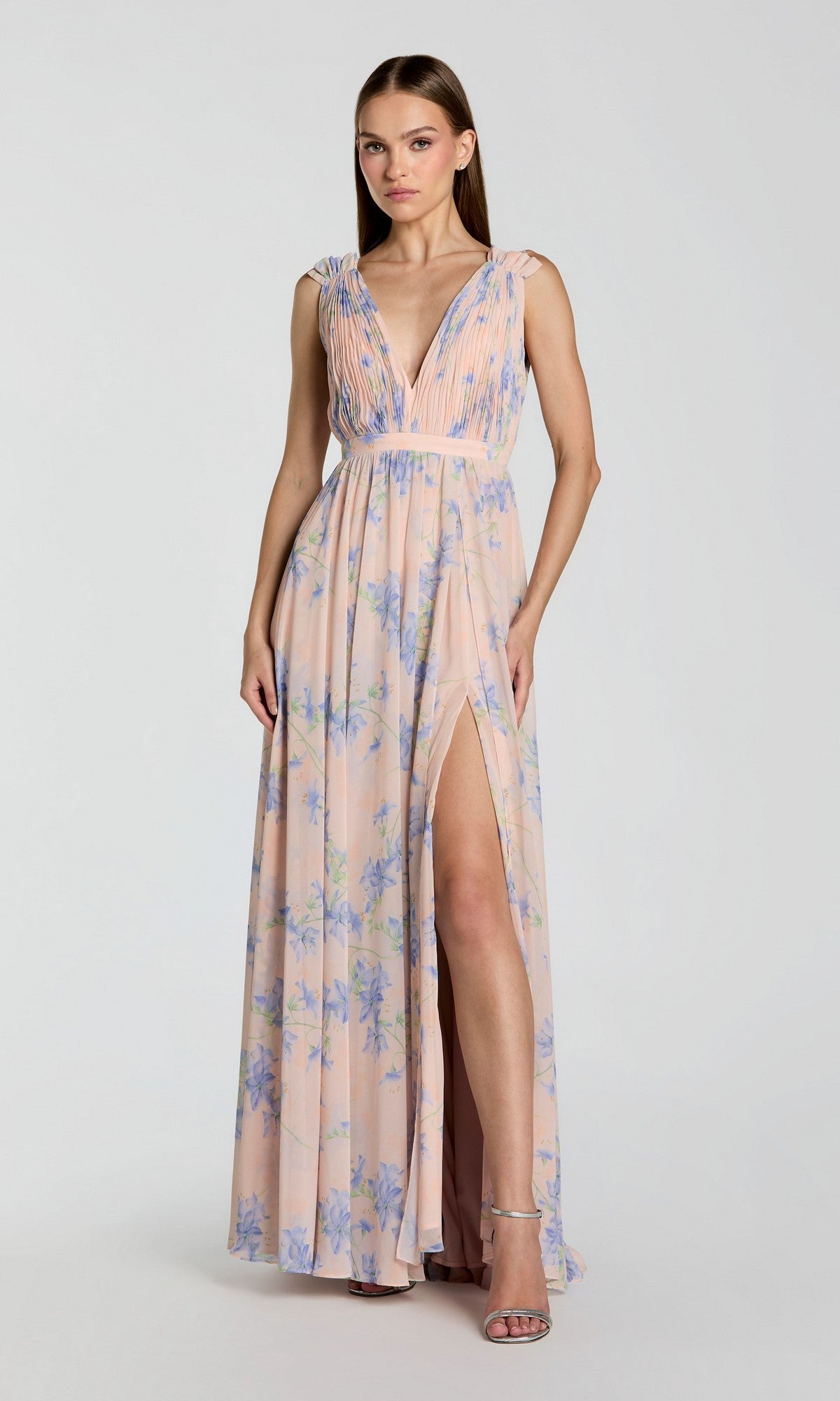 A woman wears a sleeveless, floor-length violet multi Mac Duggal 55321 evening gown with blue floral print, deep V-neckline, pleated bodice, and thigh-high slit as she stands elegantly against a plain background.