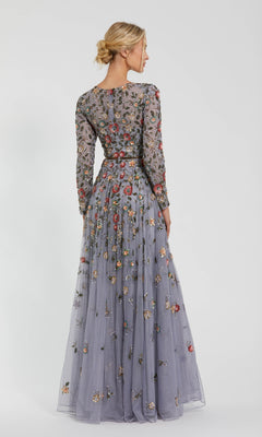 A woman stands facing away, wearing the Mac Duggal 5533 Beaded Long Sleeve Formal Dress, featuring sheer lavender fabric, colorful floral embroidery, and a flowing A-line silhouette.