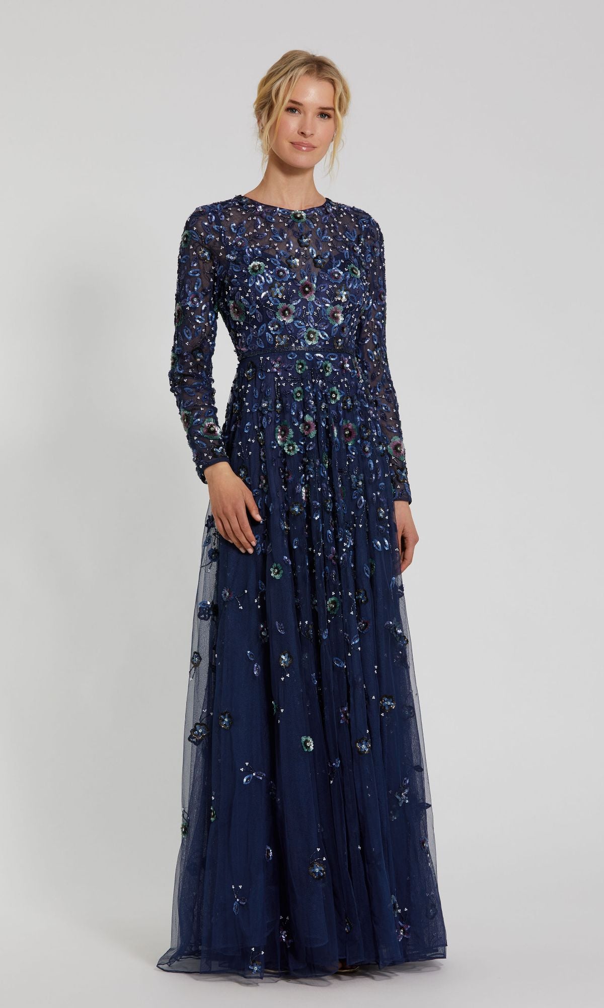 A woman wears the Mac Duggal 5533 Beaded Long Sleeve Formal Dress, a floor-length navy blue gown with floral embroidery, set against a plain light background.