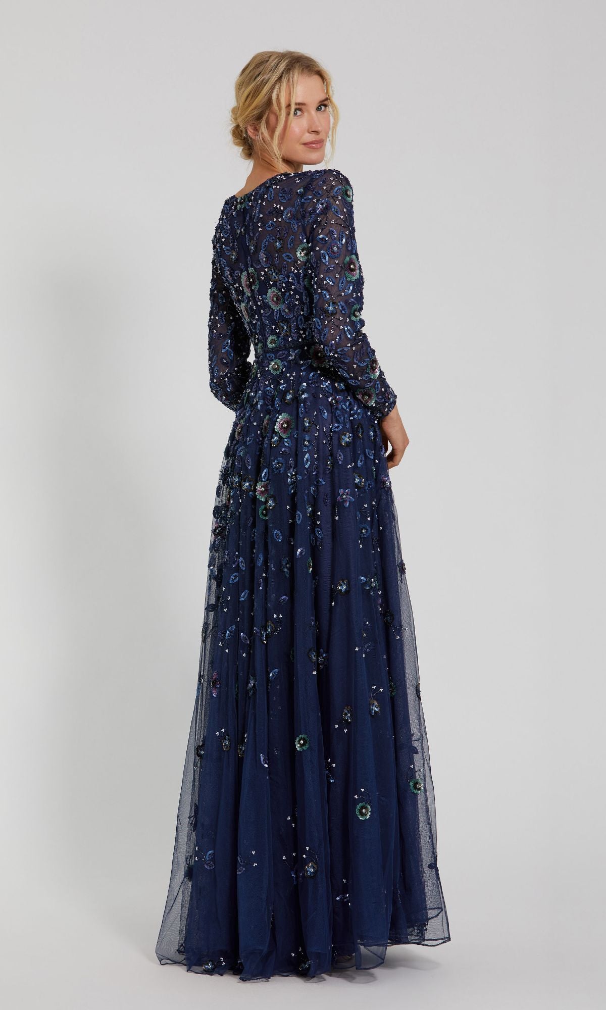 A woman stands facing away, wearing the Mac Duggal 5533 Beaded Long Sleeve Formal Dress—a floor-length navy blue gown detailed with floral embroidery and intricate beading.
