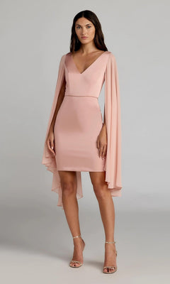 A woman stands against a plain background in the Mac Duggal 55358 short semi-formal dress in blush, featuring a knee-length V-neck design with long flowing chiffon cape sleeves and gold open-toe heels.