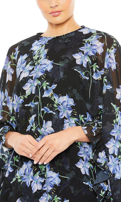 A woman models the Mac Duggal 55404 Short Print Cape Hoco Dress, featuring a black base with blue and purple floral patterns, shown from shoulders to mid-torso with hands clasped in front.