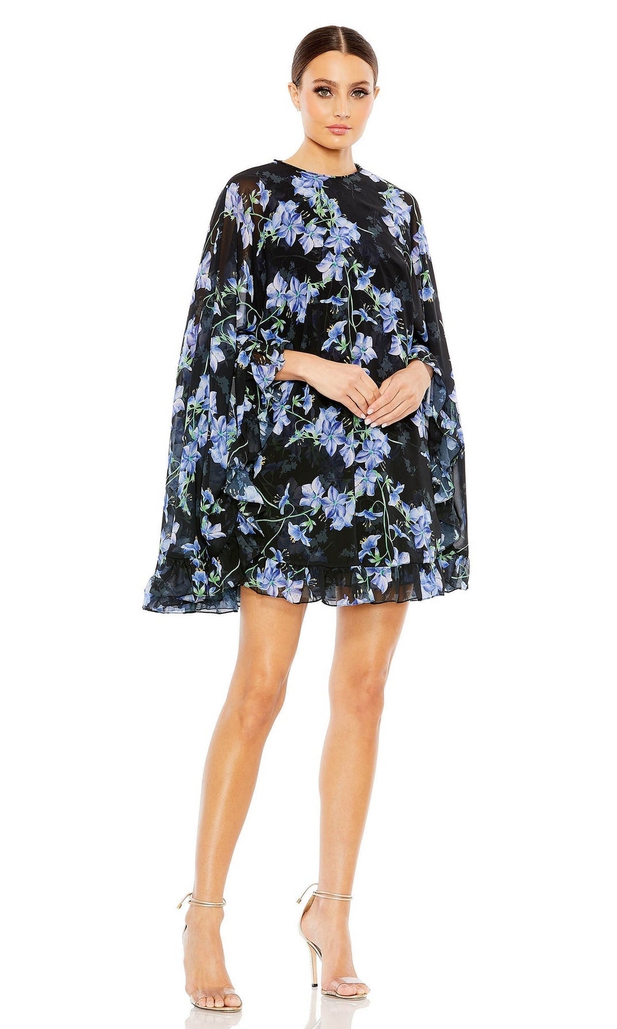 A woman models the Mac Duggal 55404 Short Print Cape Hoco Dress, featuring a loose black mini silhouette with blue floral prints and long cape sleeves, styled with silver high-heeled sandals against a white background.