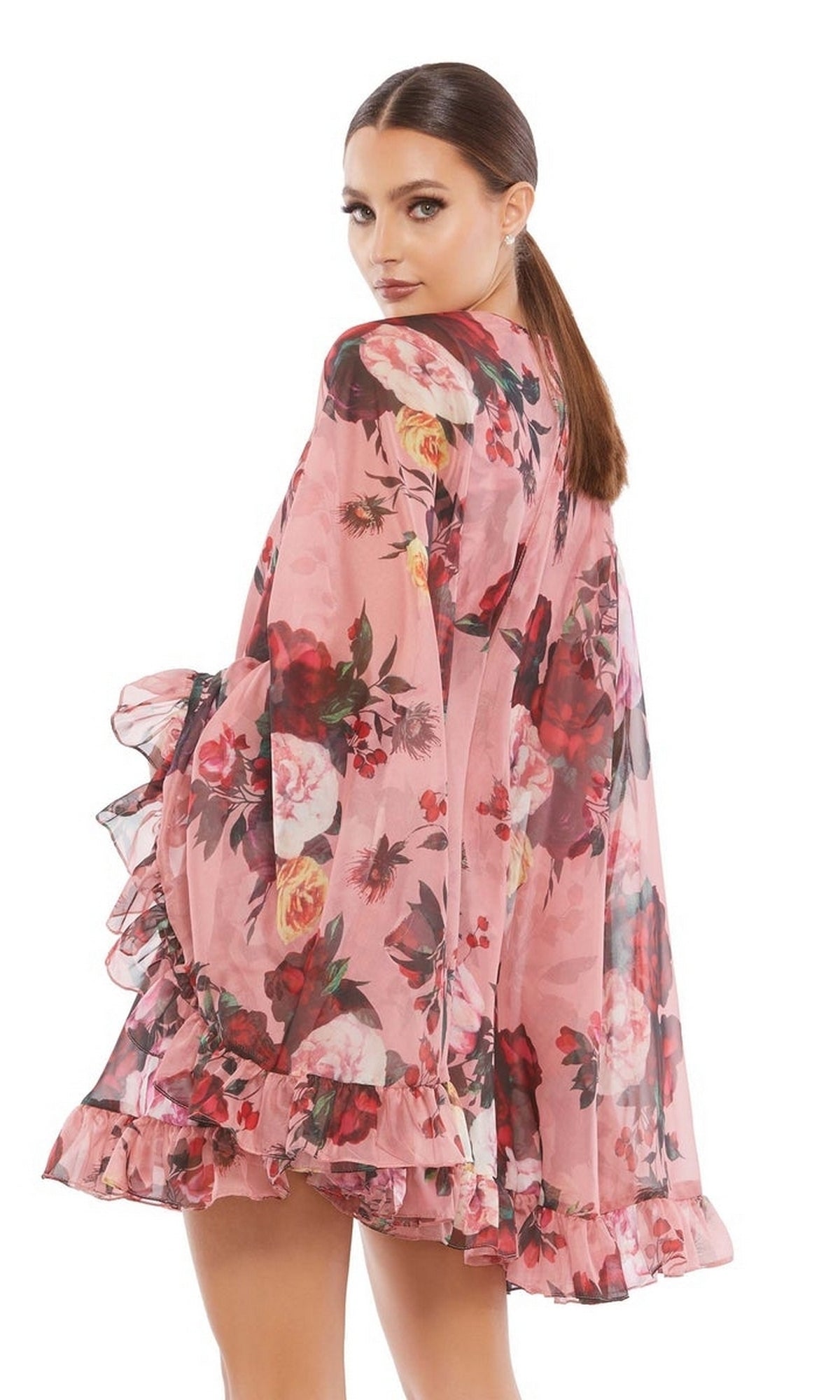 A woman stands looking over her shoulder in a pink Mac Duggal 55404 Short Print Cape Hoco Dress with floral print, long sleeves, and ruffles, set against a plain white background.