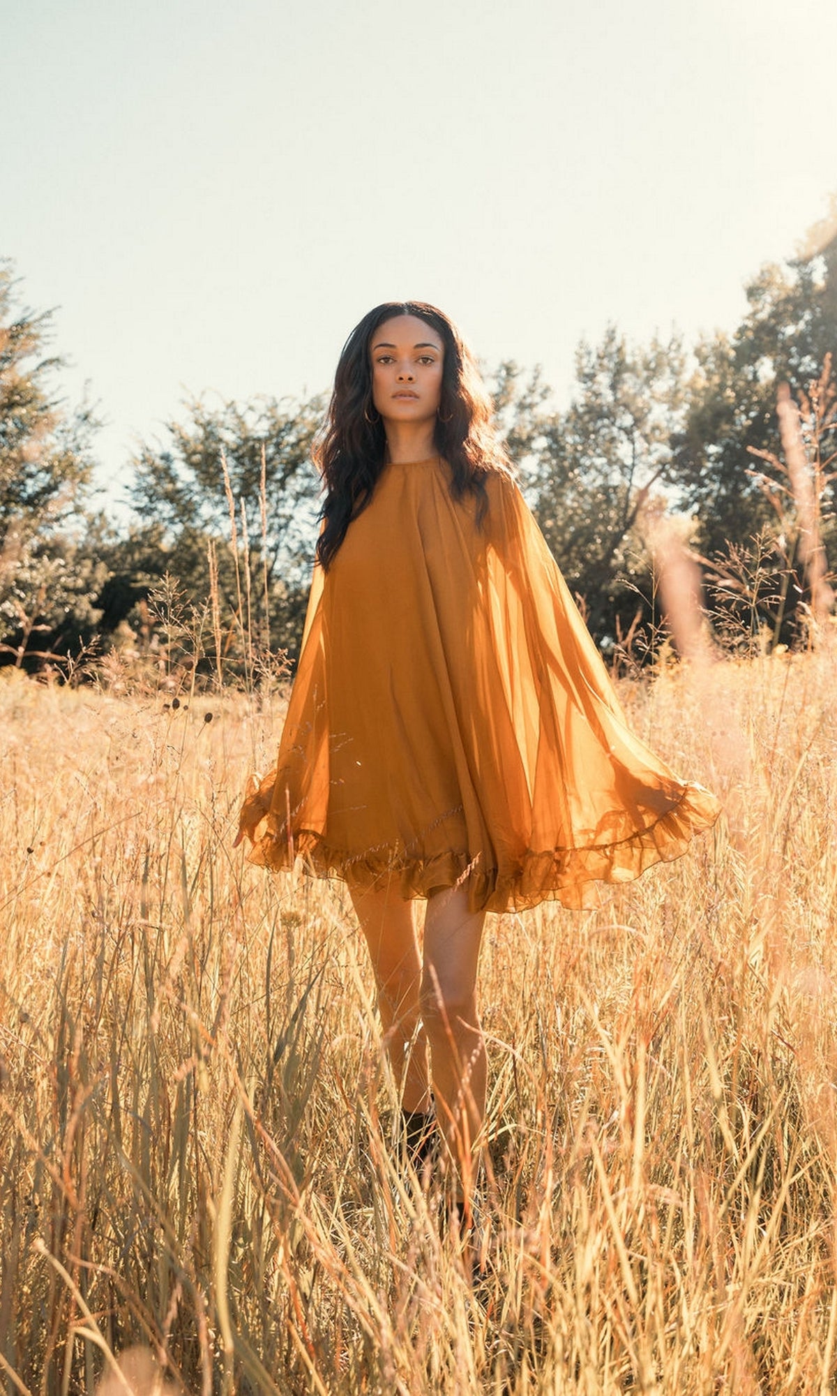 In a sunlit field of tall, golden grass with trees behind, someone wears the Mac Duggal 55407 Cape-Sleeve Short Ruffled Party Dress in orange, creating a radiant, semi-formal look.