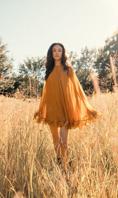 In a sunlit field of tall, golden grass with trees behind, someone wears the Mac Duggal 55407 Cape-Sleeve Short Ruffled Party Dress in orange, creating a radiant, semi-formal look.