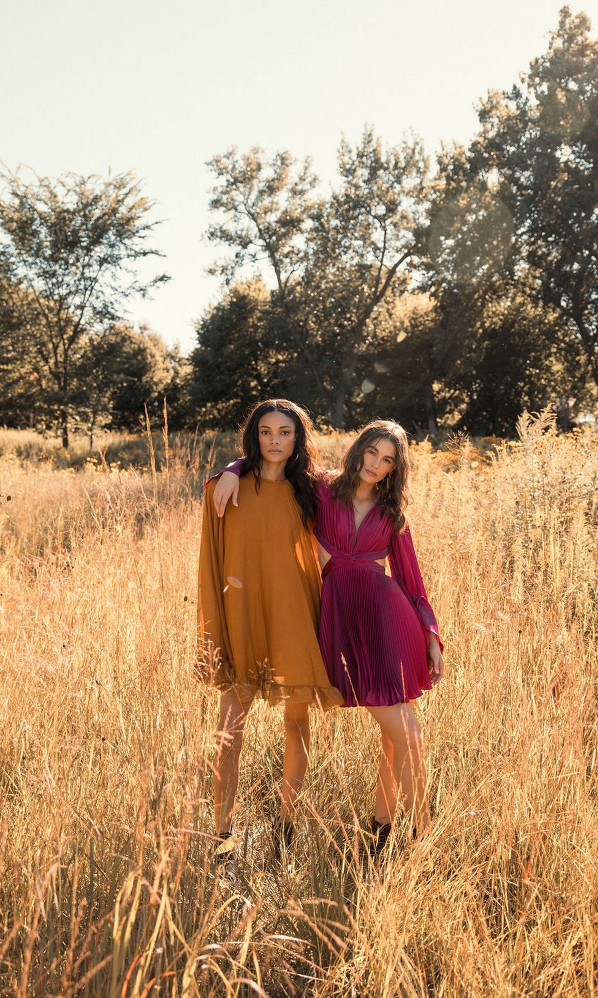 Two women stand in a sunlit field with tall grass, one wearing the Mac Duggal 55407 Cape-Sleeve Short Ruffled Party Dress in mustard yellow and the other in a magenta semi-formal cocktail dress, framed by trees.