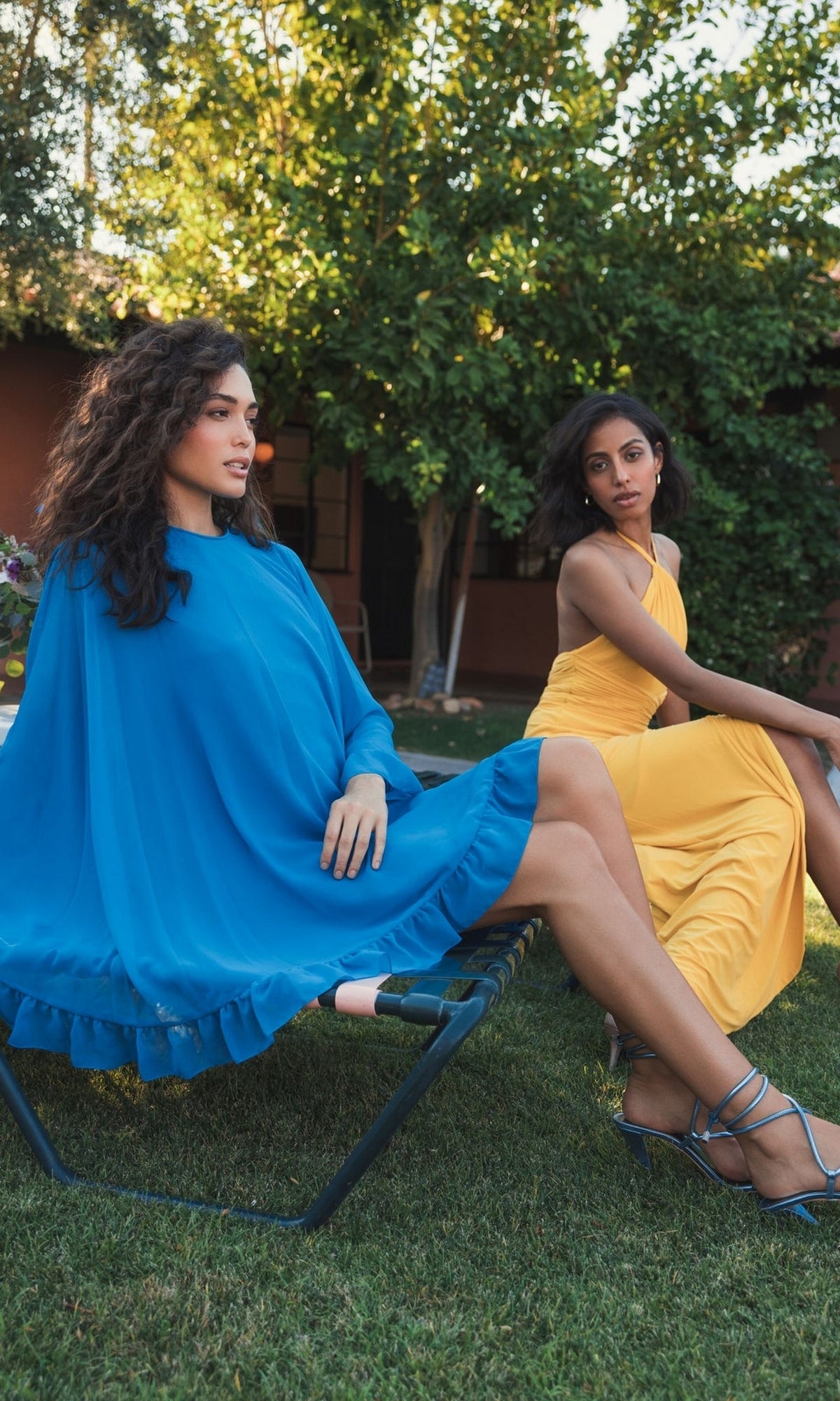 Two women sit outdoors in lawn chairs; one wears the Mac Duggal 55407 Cape-Sleeve Short Ruffled Party Dress in blue with matching heels, while the other is dressed in a fitted yellow cocktail dress and sandals. Trees and a house are visible behind them.