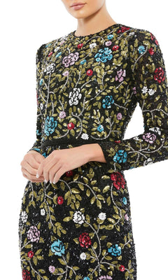 A woman stands with hands clasped in the Mac Duggal 5556—an elegant long black print formal dress adorned with colorful embroidery and sequins, perfect for any special occasion.