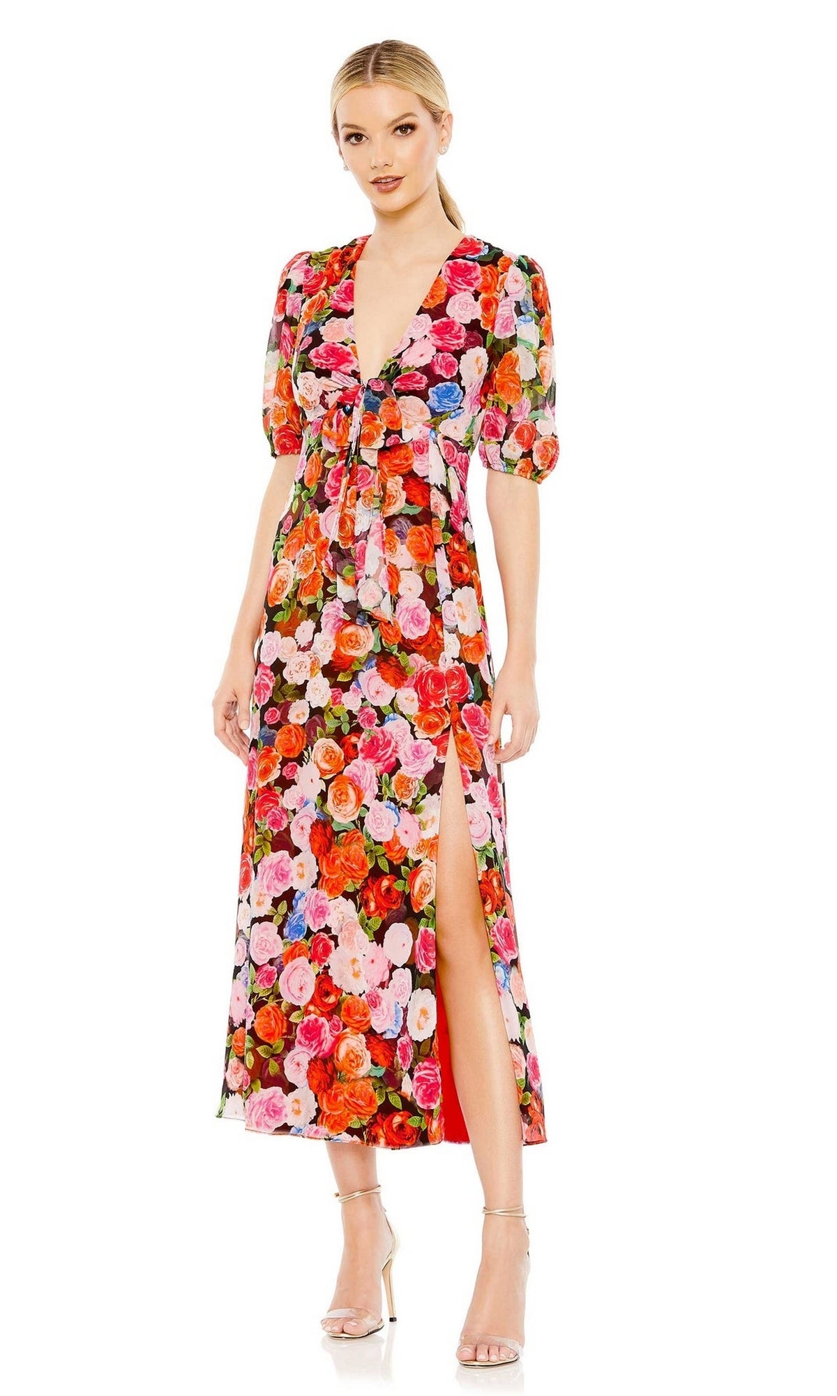 A woman wears the Mac Duggal 55626 Short Sleeve Midi Print Party Dress, featuring a vibrant floral print, puff sleeves, deep V-neckline, and thigh-high slit, styled with clear heels for a chic party look.