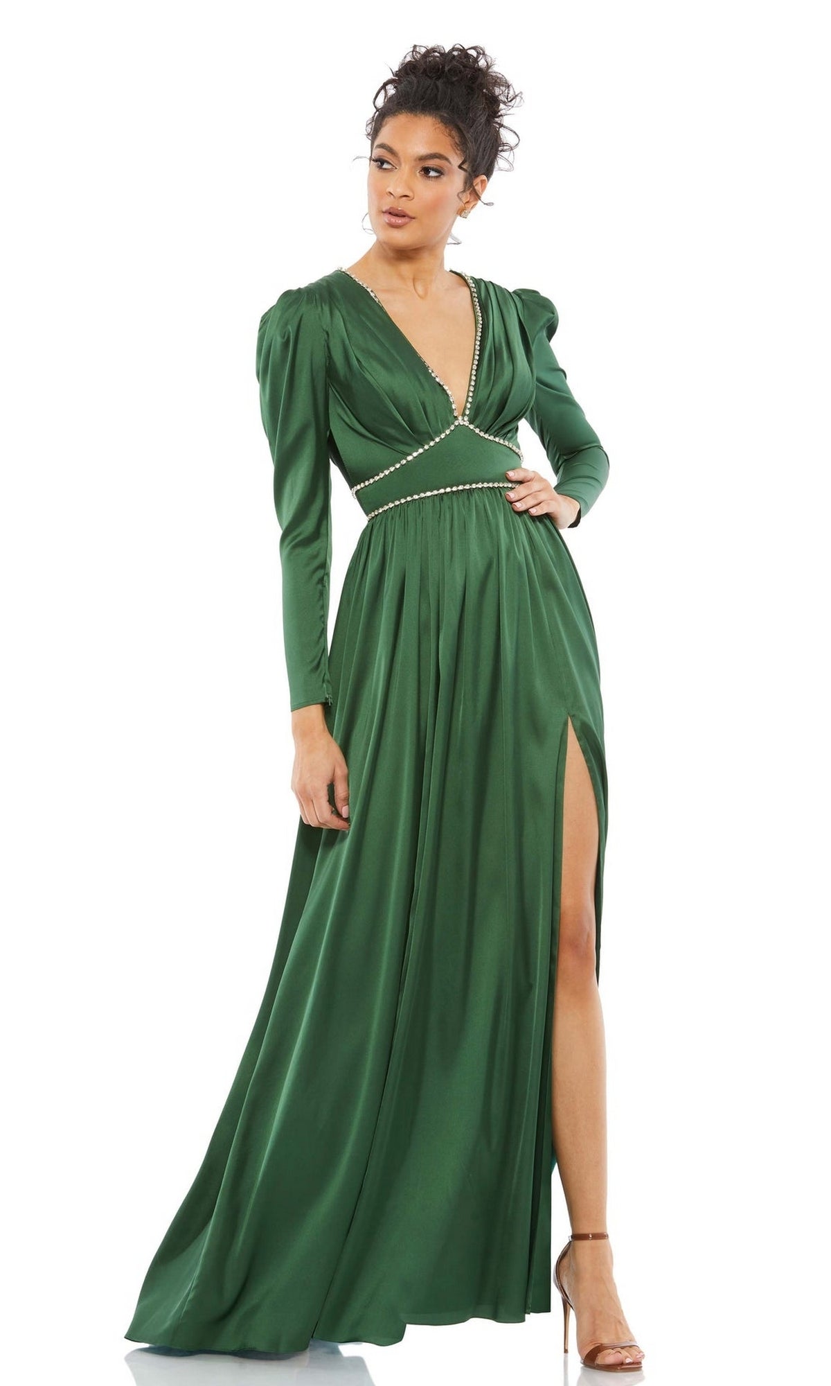 A woman wears the Mac Duggal Long Formal Dress 55702 in emerald green, featuring long sleeves, a deep V-neckline, pearl waist detail, rhinestone trim, and a high side slit, paired with strappy high-heeled sandals.