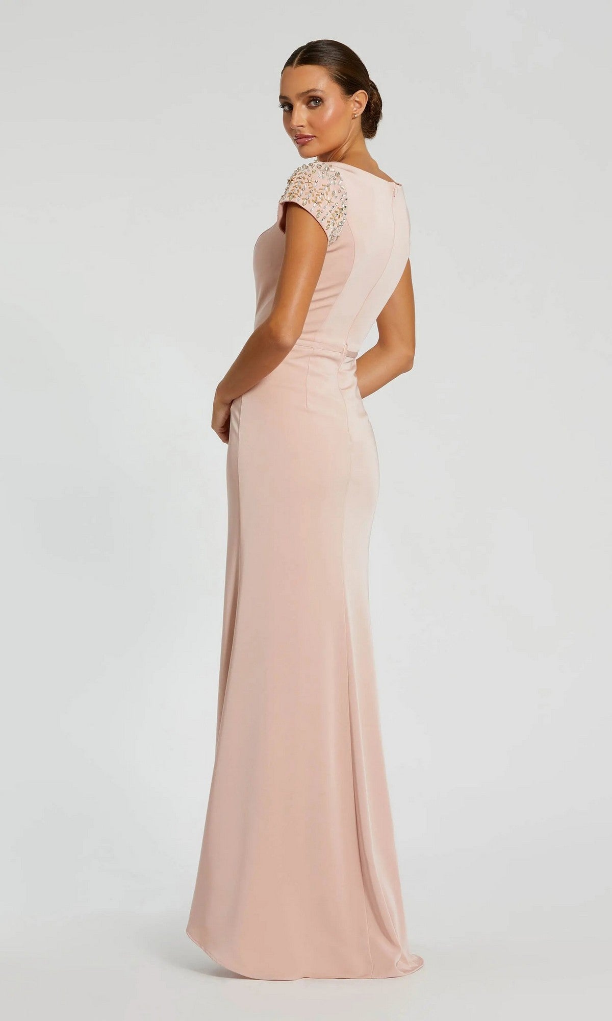 A woman stands in profile against a plain background, showcasing the Long Formal Dress 55718 by Mac Duggal—a blush pink, cap sleeve, floor-length gown with embellishments that exudes elegance and grace.