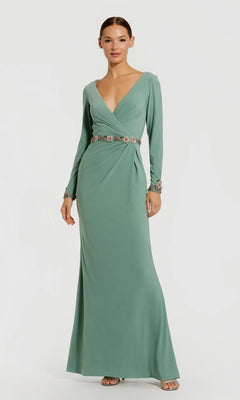A woman stands wearing the Mac Duggal 55784 long formal dress in sage, featuring long sleeves, a V-neckline, and a floral beaded waistband that adds delicate detail at the waist and cuffs.