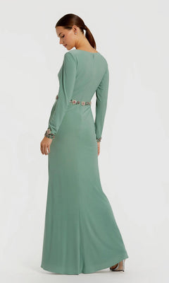 A woman stands sideways and looks down, wearing the Mac Duggal 55784 long formal dress—an elegant floor-length, sage green gown with long sleeves, a floral beaded waistband, and embellished cuffs.