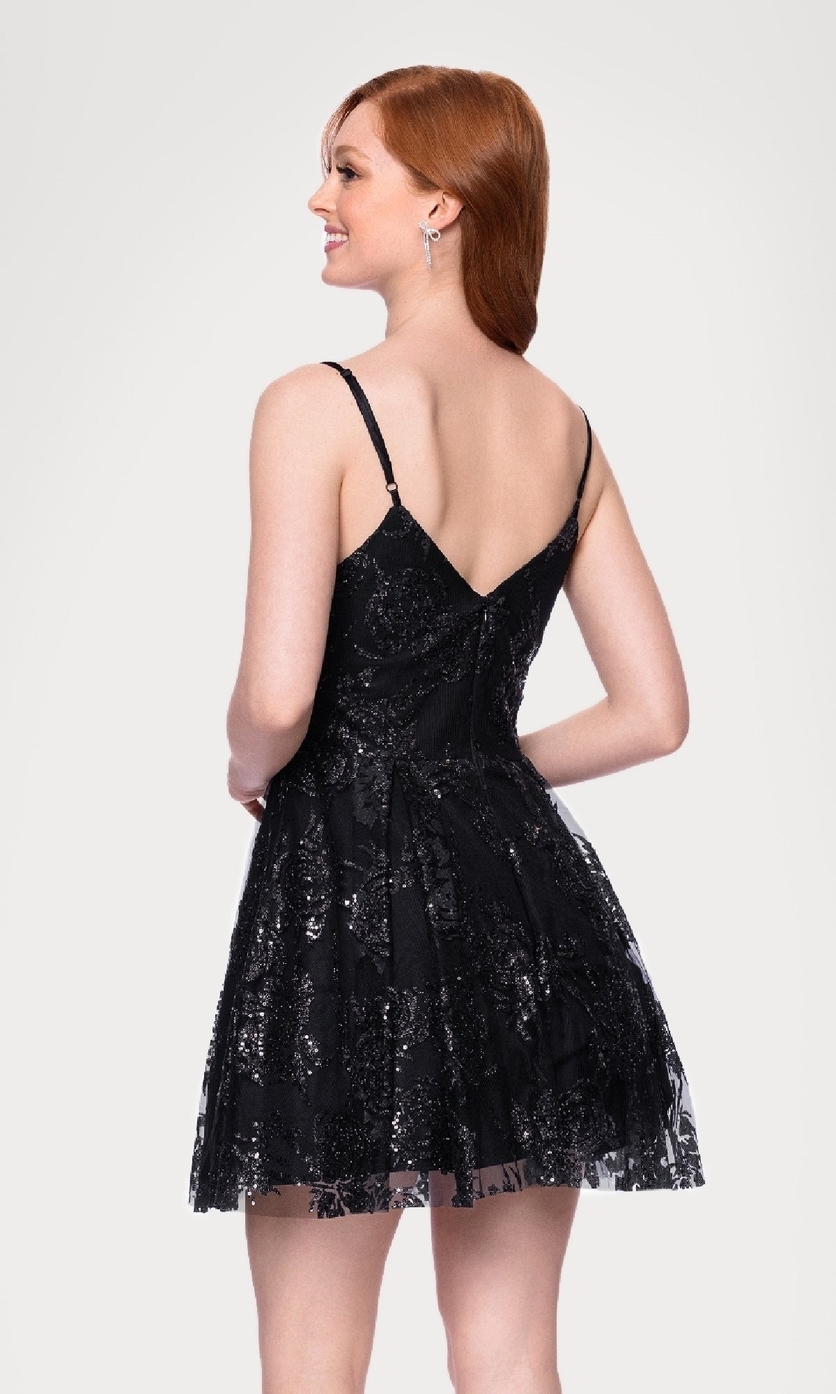 A woman with red hair smiles while looking to the side, wearing the Black Glitter Short Hoco Dress: Blondie Nites 5583BN, a junior dress featuring thin straps and black lace details.