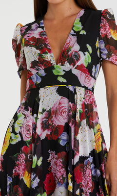 A woman wears the Mac Duggal 55933 Short Sleeve Print Formal Dress, a black V-neck style with colorful floral print, puff sleeves, and a fitted waist.