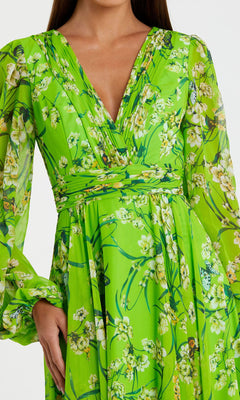 A woman wears the Long Green Print Formal Dress: Mac Duggal 56013, featuring a bright green floral print, long sleeves, and a V-neckline; her left hand rests on her hip.