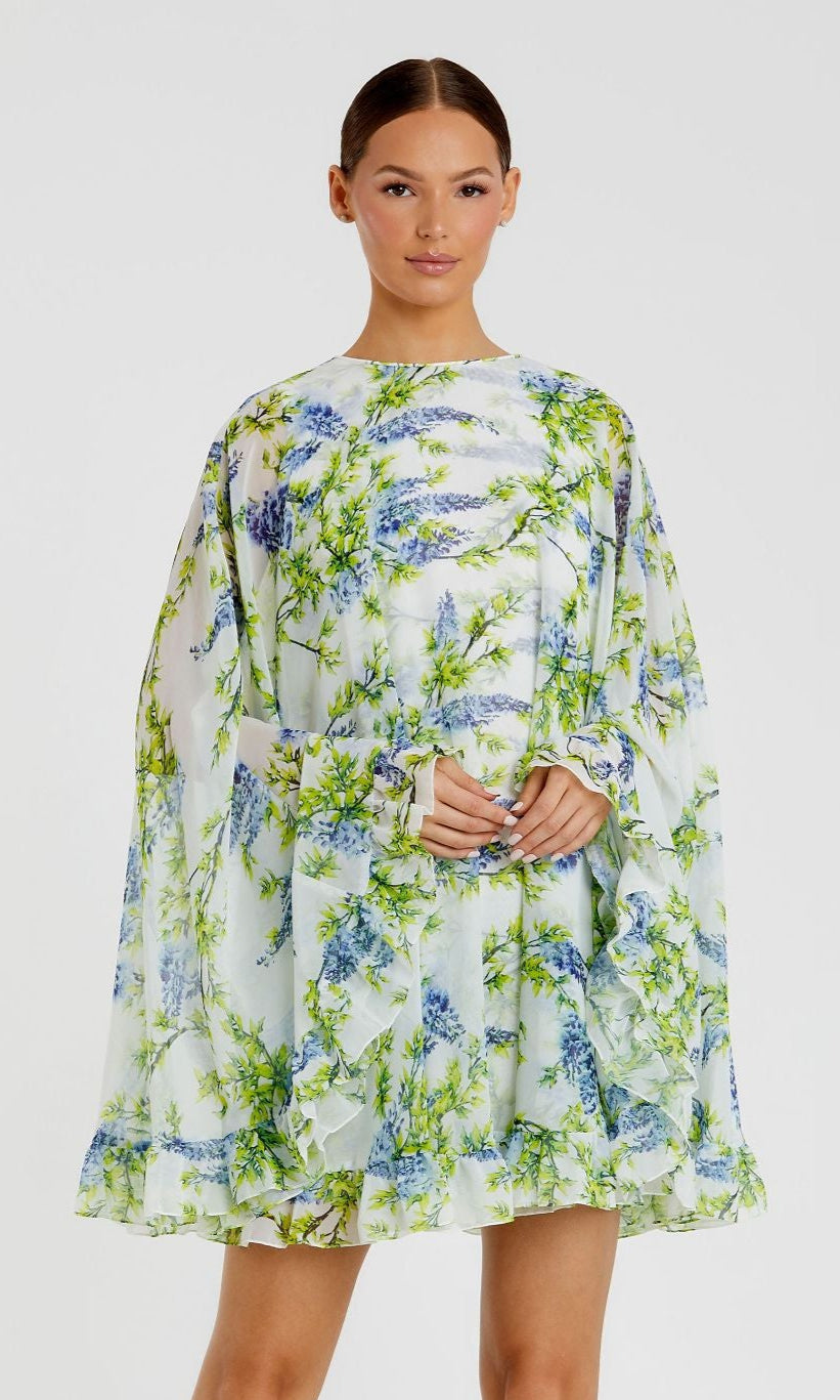 A woman models the Cape Short Party Dress 56029 by Mac Duggal, a white floral party dress with long sleeves and a blue-green pattern, standing against a plain white background.