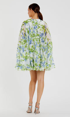 A woman faces away, wearing the Mac Duggal Cape Short Party Dress 56029—a loose, short white floral dress with green and blue patterns—and silver high-heeled sandals against a plain white background.