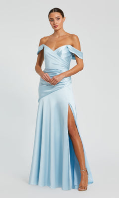A woman in the Mac Duggal 56037 off-the-shoulder prom dress, featuring light blue satin and a thigh-high slit, pairs it with strappy heels while standing against a plain white background.