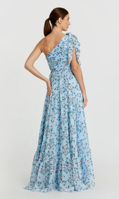 A woman in a light blue, floral, floor-length Mac Duggal 56051 formal dress stands with her back to the camera, hair tied in a low ponytail.