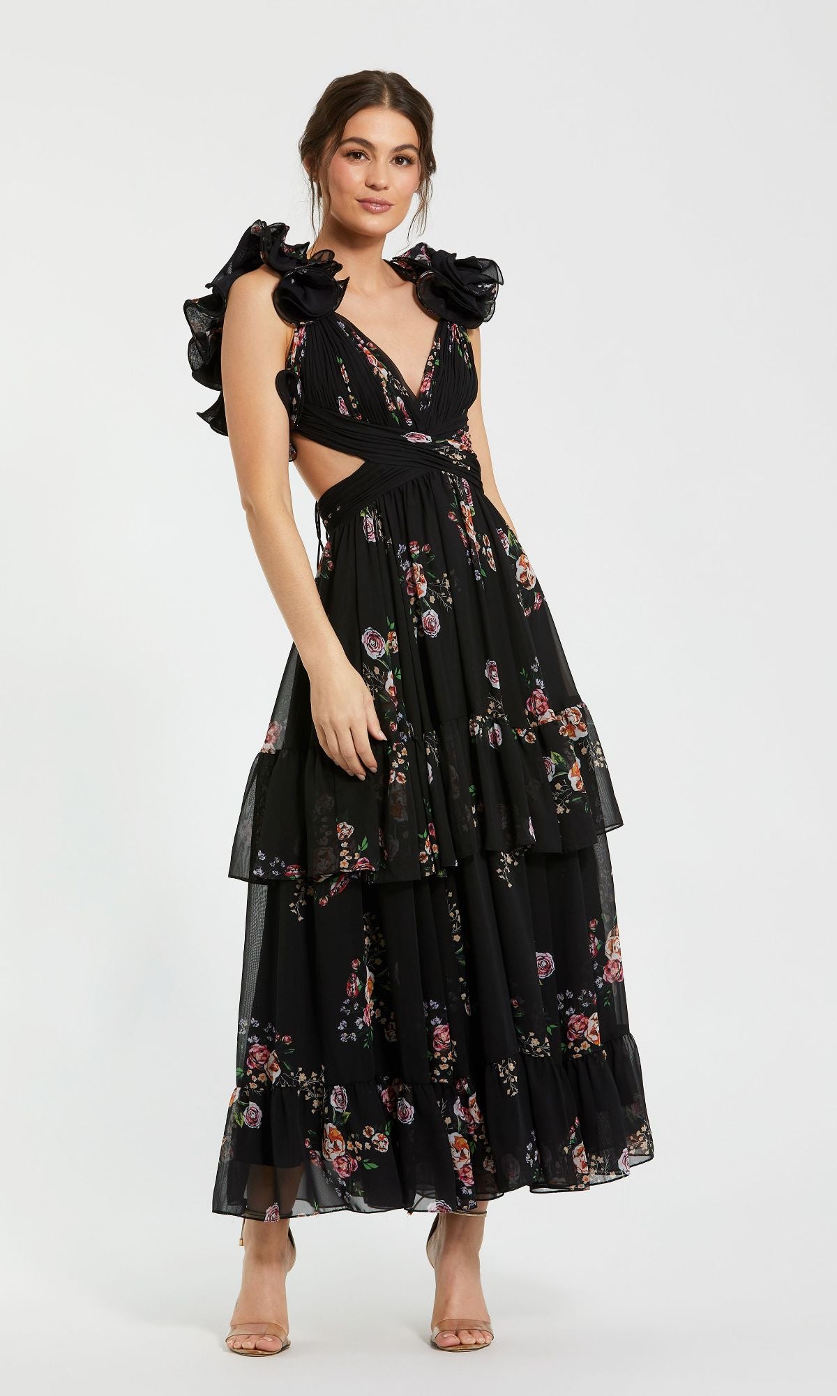 A woman models the Mac Duggal 56054 Cut-Out Black Print Formal Dress, featuring a long floral design, side cutouts, and ruffled sleeves, standing against a plain white background.