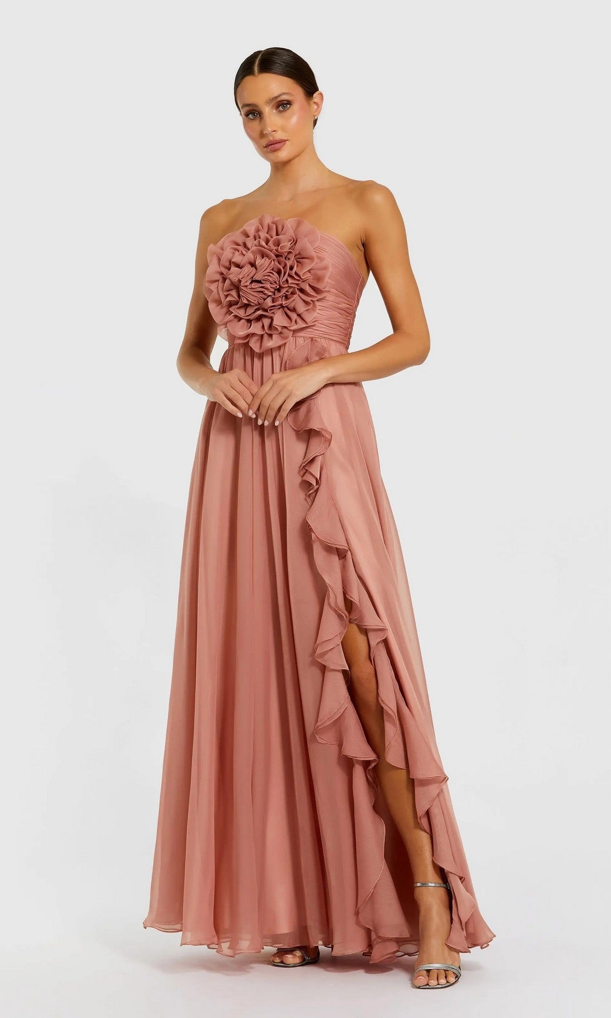 A woman stands against a plain white background wearing the Mac Duggal 56071 Long Formal Dress in Rose—an elegant strapless chiffon gown with a dramatic 3D floral bodice, ruffled slit, and paired with high-heeled sandals.