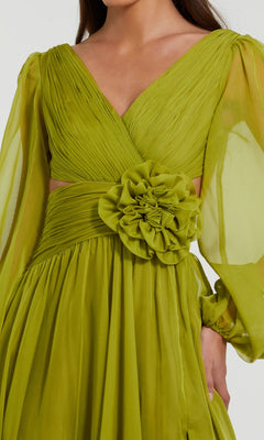 A woman wearing the Mac Duggal 56072 long formal dress, featuring light green chiffon fabric, long bishop sleeves, a deep V-neckline, ruched bodice, a large fabric flower at the waist, and an elegant open back design.