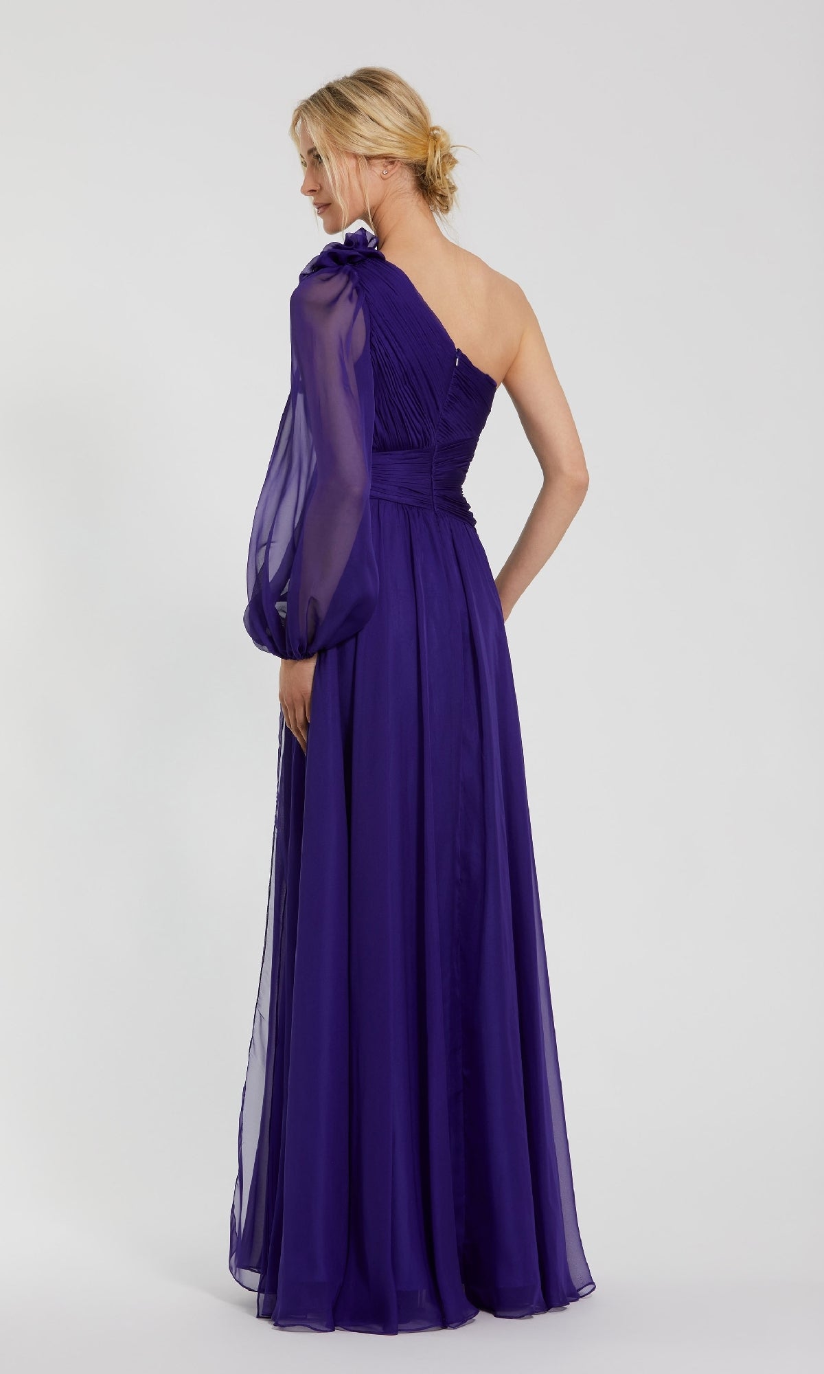 A woman stands facing away against a plain light background, wearing the Mac Duggal 56073: a floor-length, one-shoulder purple formal dress with a sheer long sleeve.