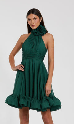 A woman wears the Short Wedding Guest Dress 56080 by Mac Duggal, a sleeveless teal green dress with a halter neckline, large ruffle accent, and ruffled hem, standing against a plain light background.