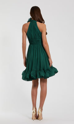 A woman stands facing away in the Short Wedding Guest Dress 56080 by Mac Duggal, a sleeveless teal green dress with a ruffled hem, paired with gold high-heeled shoes against a plain light background.
