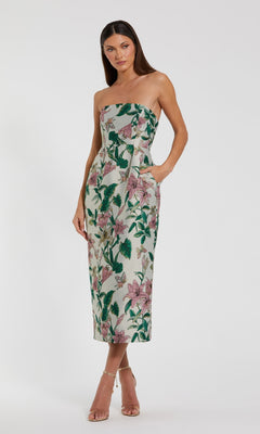 A woman stands wearing the Mac Duggal 56097 Midi Floral Wedding Guest Dress, a strapless midi dress with green and pink floral accents, paired with light high-heeled sandals against a plain white background.