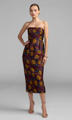 A woman in the Mac Duggal 56097 Midi Floral Wedding Guest Dress in Plum Multi stands against a plain gray background, paired with gold sandals and minimal jewelry—an elegant choice for a wedding guest ensemble.