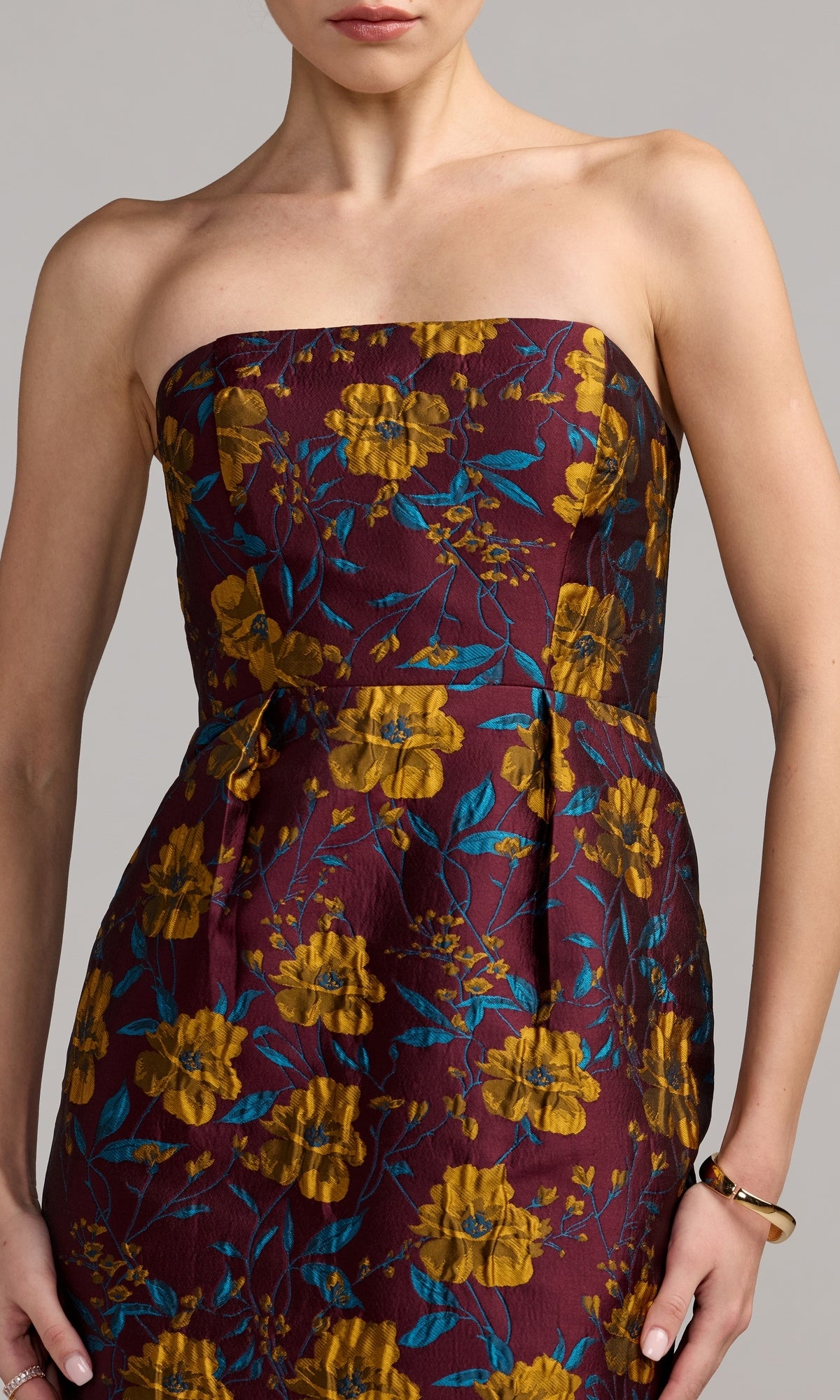 A woman is wearing the Mac Duggal 56097 Midi Floral Wedding Guest Dress—a strapless, maroon-colored dress adorned with yellow floral and blue leaf patterns. Only her upper body is visible against a neutral background.