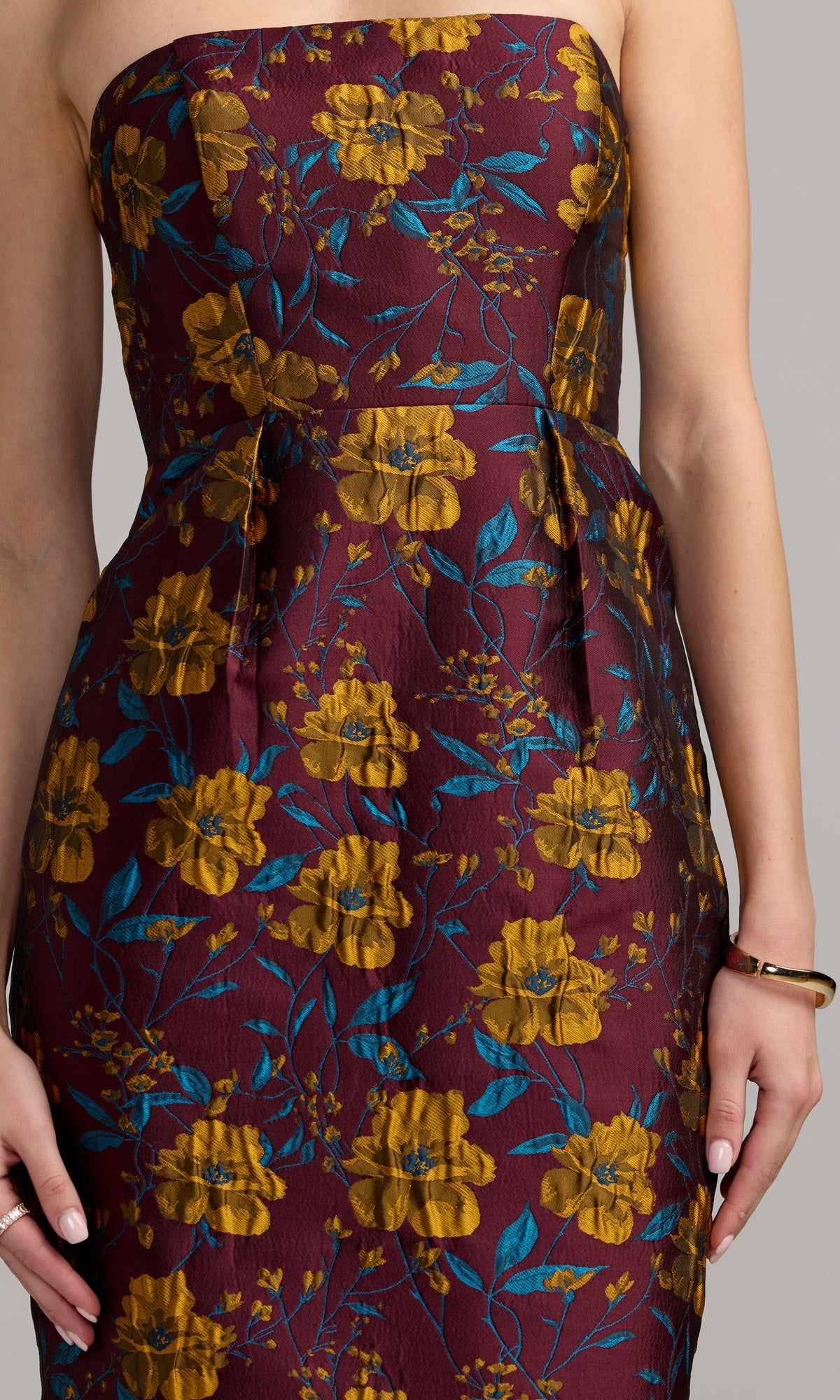 A woman wearing the Midi Floral Wedding Guest Dress: Mac Duggal 56097—a strapless maroon tea-length party dress featuring a yellow floral and teal leaf pattern—stands with her arms relaxed by her sides.
