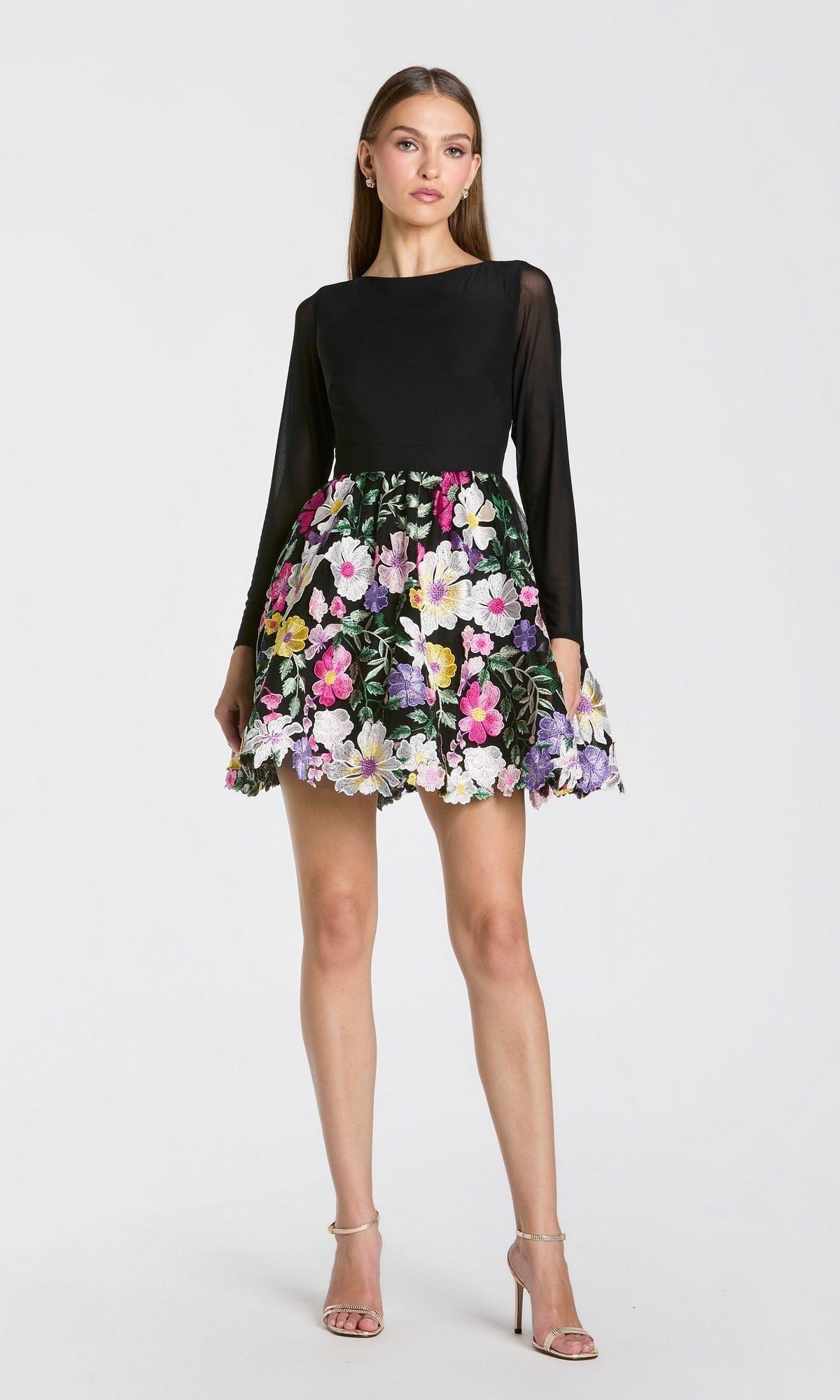 A woman stands against a plain white background wearing the Mac Duggal 56109 short party dress in Black Multi, featuring long sleeves and a vibrant, floral-embroidered skirt, paired with strappy high heels.