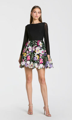 A woman stands against a plain white background wearing the Mac Duggal 56109 short party dress in Black Multi, featuring long sleeves and a vibrant, floral-embroidered skirt, paired with strappy high heels.