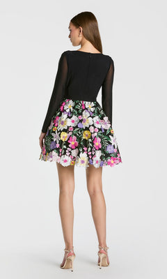 A woman faces away, modeling the Mac Duggal 56109 short party dress—a black long-sleeve style with a flared, floral-embroidered skirt—paired with nude high-heeled sandals against a plain white background.