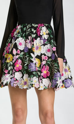 Wearing a black long-sleeve top with the Mac Duggal 56109 short party dress—a skirt beautifully detailed with colorful floral embroidery in pink, purple, yellow, and white.