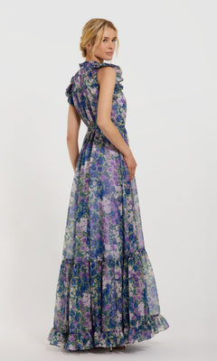 A woman models the Mac Duggal 56116 Lilac Floral-Print Formal Gown, a long sleeveless dress in purple, blue, and green tones, standing against a plain white background and looking over her shoulder.