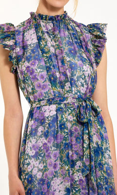 A woman wears the Lilac Floral-Print Formal Gown by Mac Duggal 56116, featuring a sleeveless, ruffled design and a matching tie belt at the waist in purple, blue, and green florals.