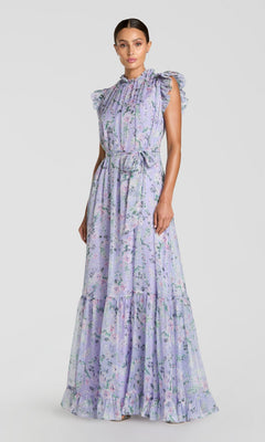 A woman stands against a plain background wearing the Mac Duggal 56116 Lilac Floral-Print Formal Gown, featuring a sleeveless, floor-length design with ruffled shoulders and a waist tie.