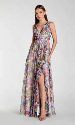 A woman stands wearing the Mac Duggal 56119 Long Prom Dress in Multi—a sleeveless, floor-length floral chiffon gown with a ruched bodice and deep V-neck, featuring an A-line skirt with a ruffled high slit—elegantly paired with strappy heels.
