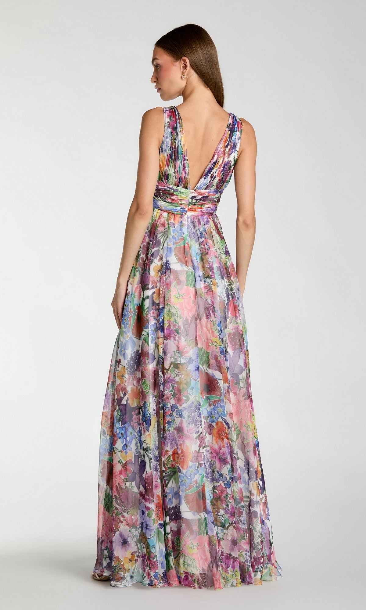 A woman stands facing away against a plain light background, wearing the Mac Duggal 56119 long prom dress. The gown features a graceful A-line skirt, a beautifully ruched sleeveless bodice, and an elegant deep V-back in soft floral chiffon.
